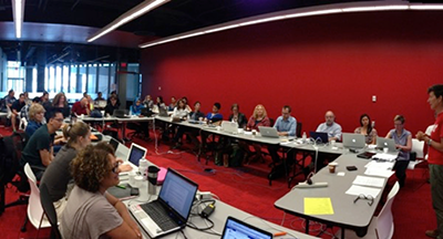 A group of people in a red room arranged in a large rectangle listening to a speaker